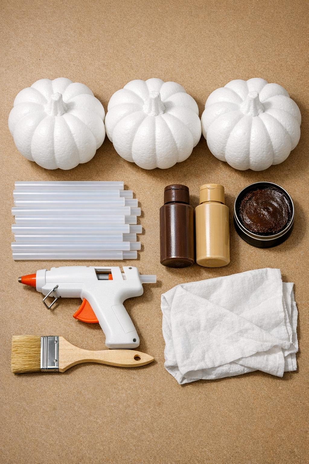 Foam pumpkins, hot glue sticks, paint, wax, glue gun, chip brush, and rag laid out on a craft table