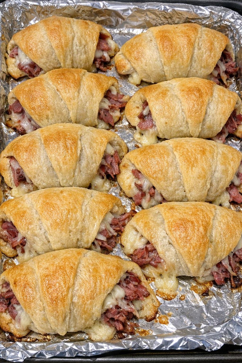 Puffy golden crescent pillows stuffed with corned beef and baby Swiss on a baking sheet