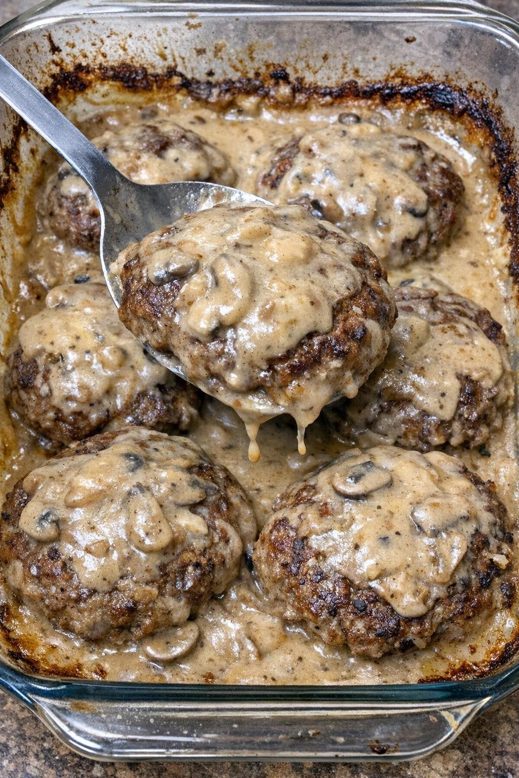 Tender browned ground beef patties smothered in creamy mushroom gravy in a glass baking dish