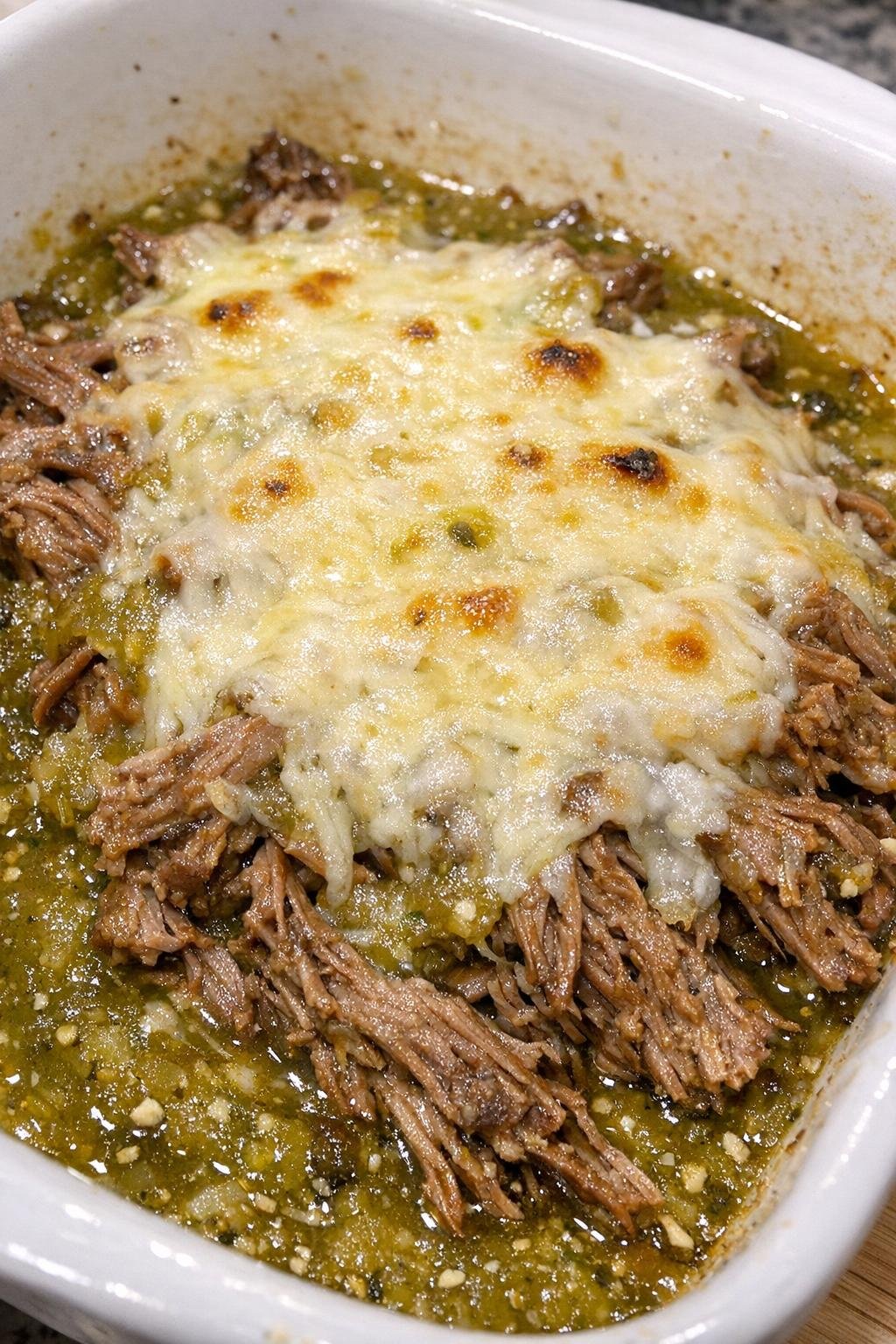 Shredded beef with salsa verde and melted cheese in a white casserole dish