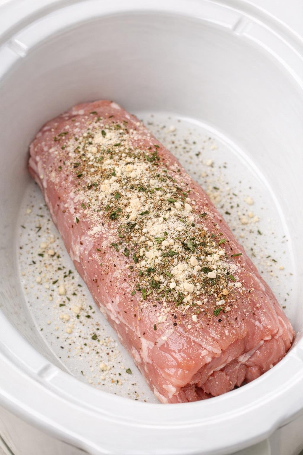 Raw pork tenderloin in a white slow cooker insert with dry seasoning