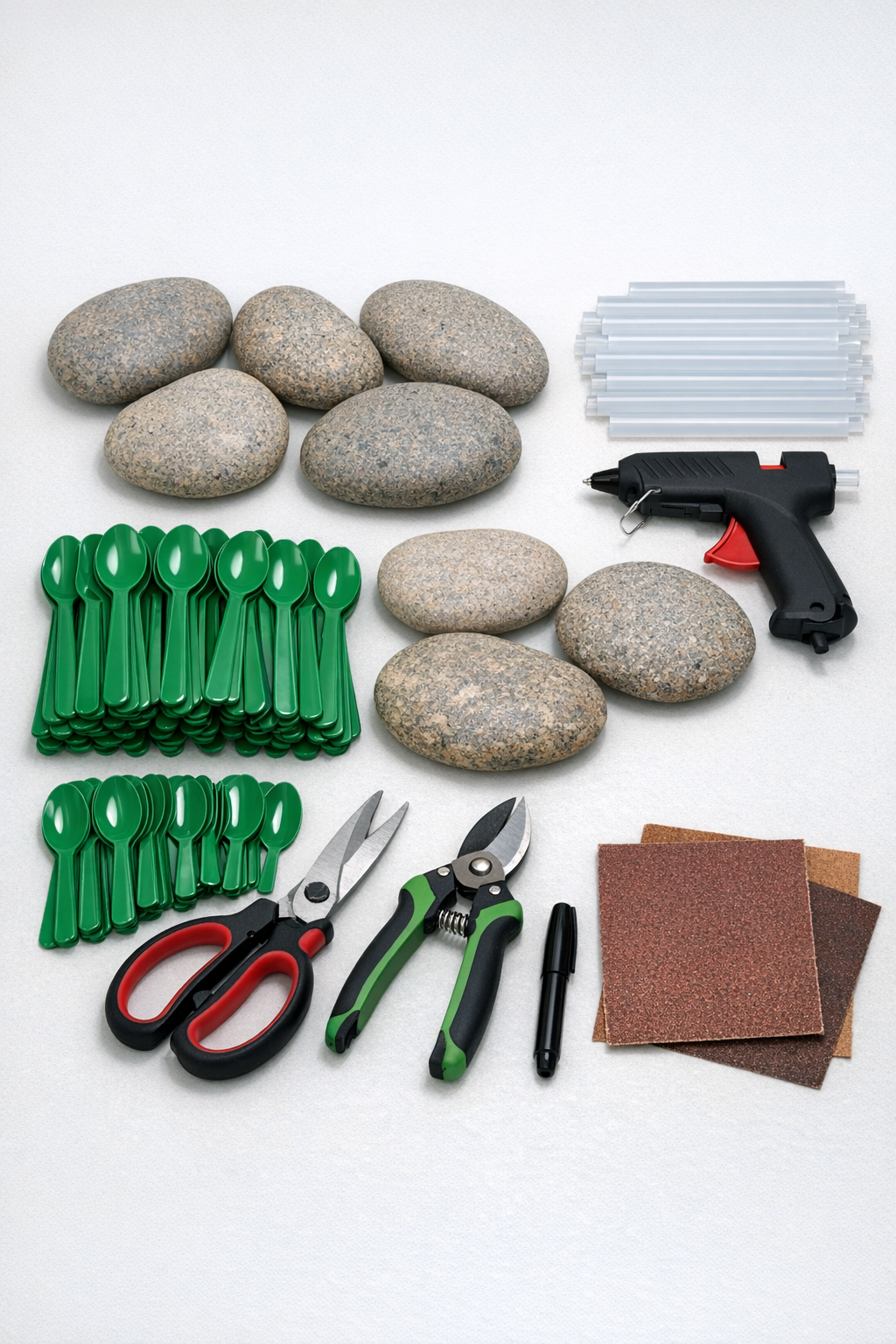Green plastic spoons, flat river rocks, glue sticks, and cutting tools laid out for the project