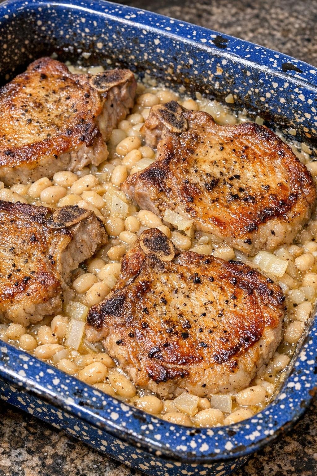 Golden browned pork chops baked over creamy navy beans in a blue speckled enamel pan