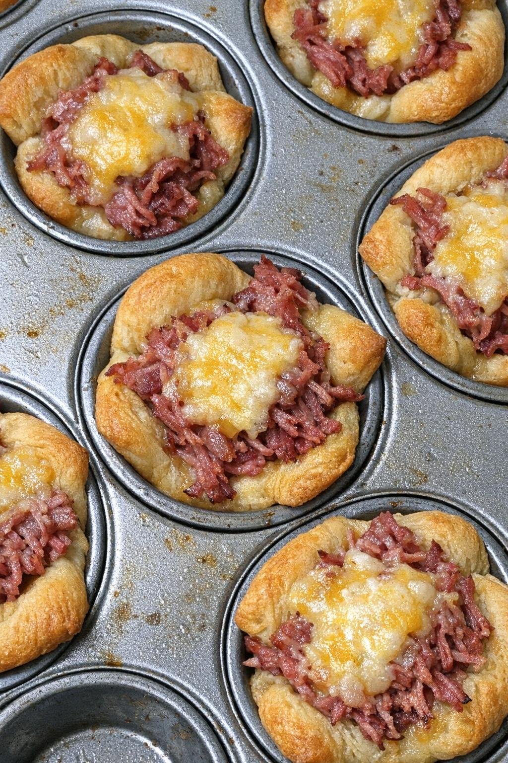 Flaky golden crescent dough cups filled with corned beef and melted cheddar in a muffin tin