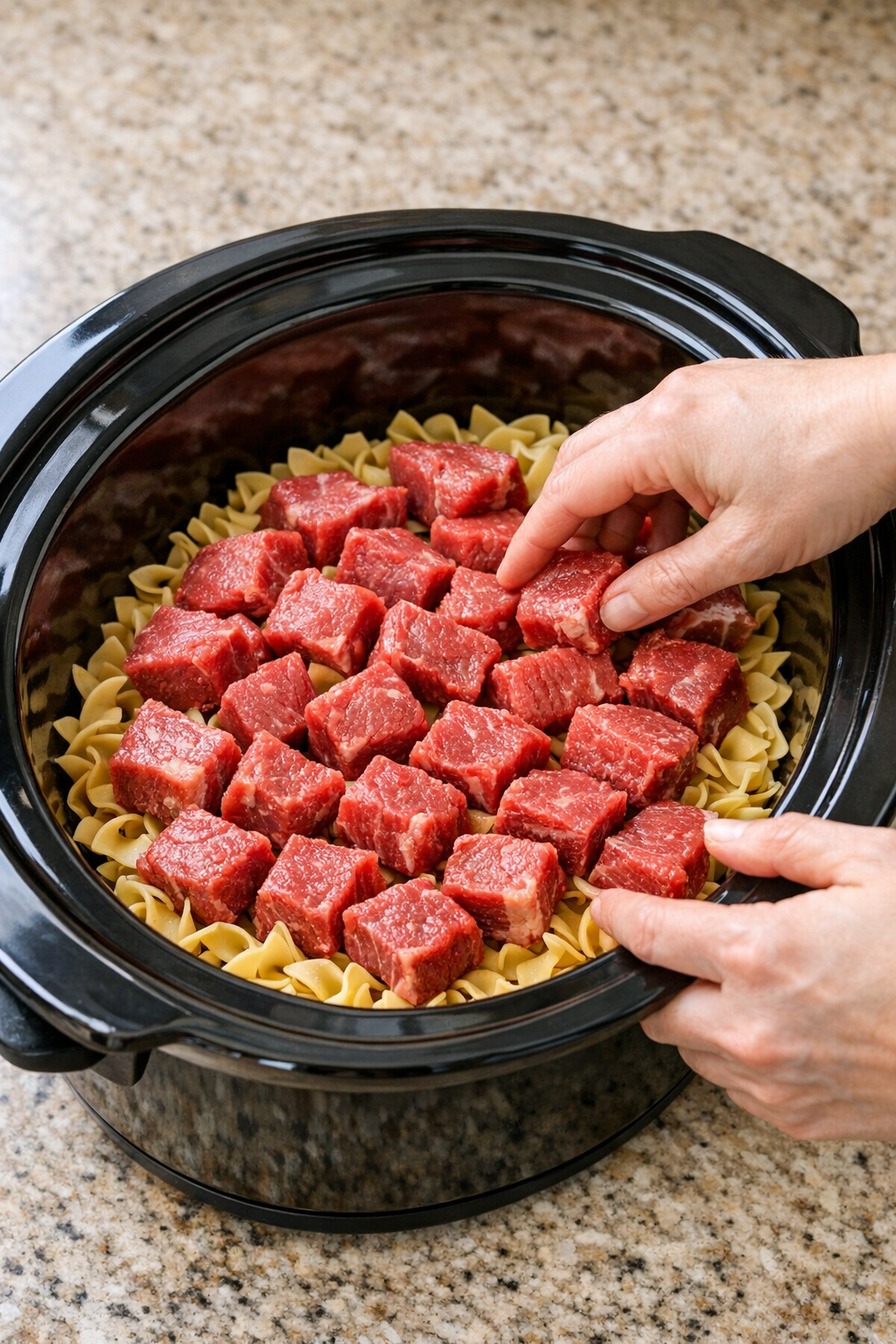 Raw beef cubes layered over egg noodles in the slow cooker