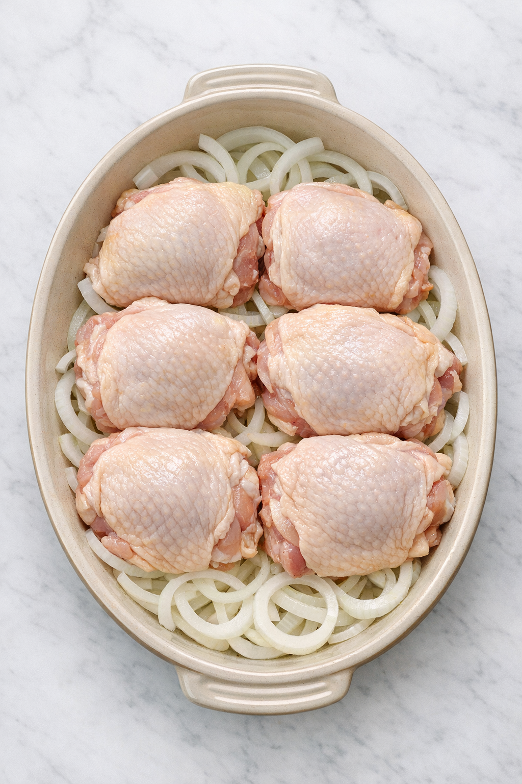 Raw chicken thighs placed over sweet onion rings in a baking dish